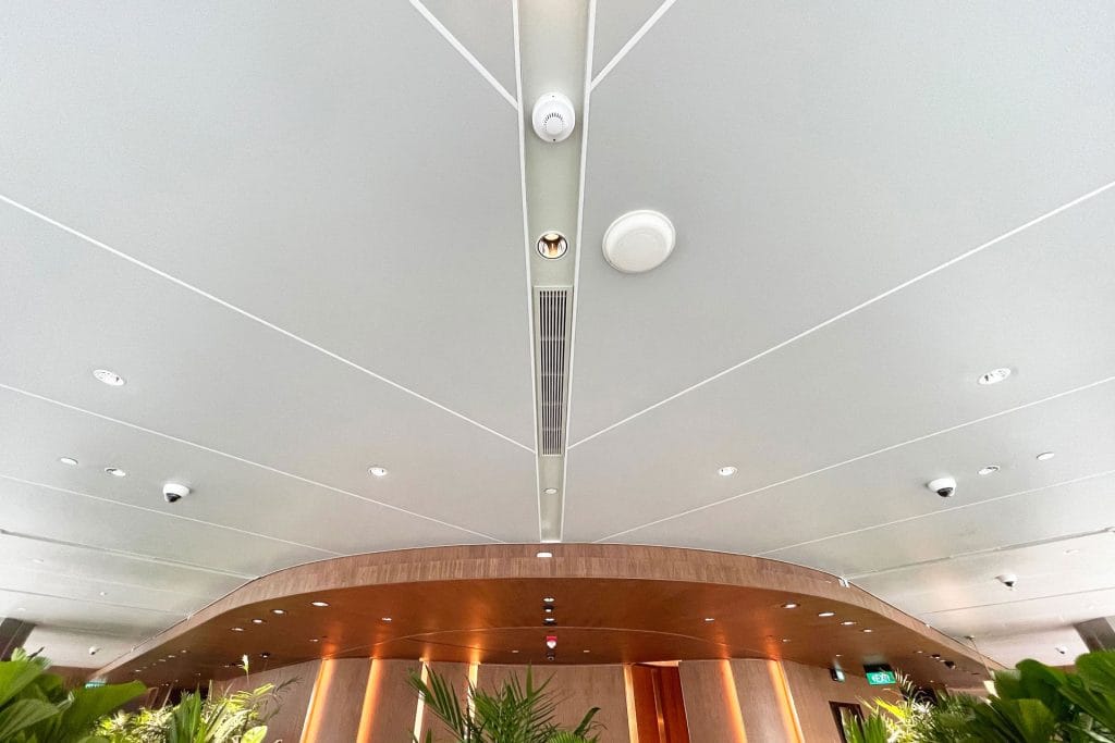 Ceiling view of a modern interior space with Ligkt™ Stretch Membrane Ceiling, recessed lighting, round smoke detectors, air vents, security cameras, and a curved wooden structure below, with green plants visible at the bottom edges.