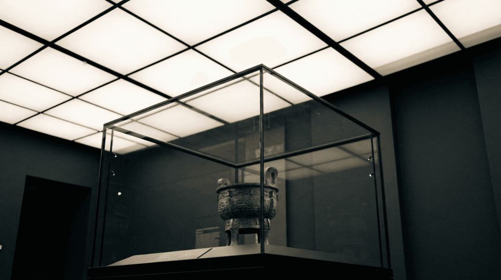 A green ancient bronze vessel is displayed inside a glass case under bright Ligkt™ Stretch Ceilings in a dimly lit museum room.