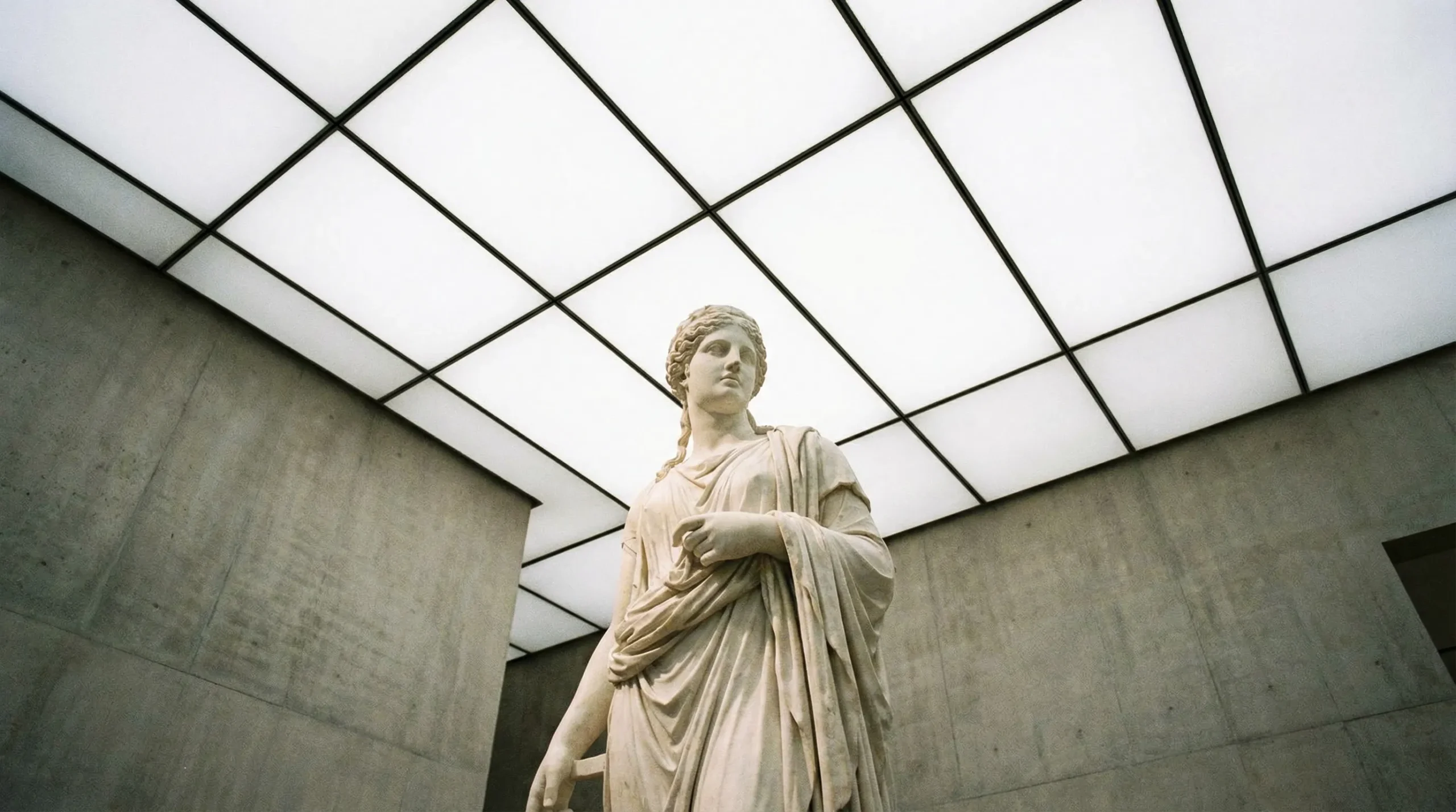 A marble statue of a woman in classical draped clothing stands in a modern, minimalist gallery with concrete walls and a large Ligkt™ Stretch Ceilings grid-pattern skylight above.