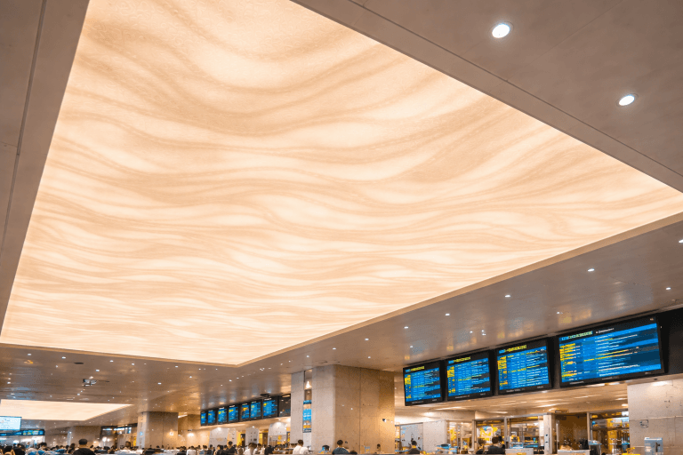 A large, illuminated Fire-Rated Stretch Ceiling with a wavy pattern spans above an airport terminal; digital flight information boards and crowds of travelers are visible below, making it ideal for busy transit hubs.