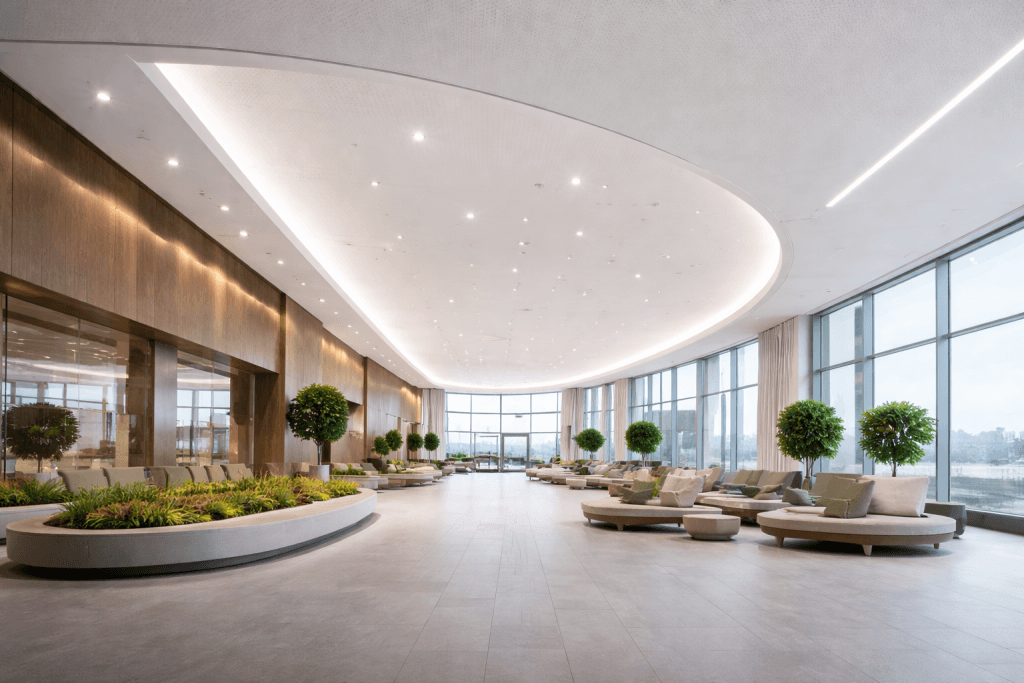A spacious, modern lounge with circular seating, green potted trees, and natural light. Curved ceiling lights—enhanced by C2C Pathways for Stretch Ceiling Systems—and lush indoor plants create a bright, inviting atmosphere.