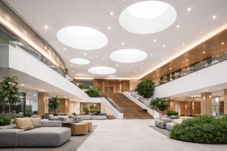 Spacious modern atrium with large circular skylights, a wide wooden staircase, indoor plants, and seating areas with gray sofas and cushions. Warm lighting complements C2C Pathways for Stretch Ceiling Systems and glass railings for an open, inviting atmosphere.