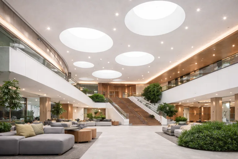 Spacious modern atrium with large circular skylights, a wide wooden staircase, indoor plants, and seating areas with gray sofas and cushions. Warm lighting complements C2C Pathways for Stretch Ceiling Systems and glass railings for an open, inviting atmosphere.