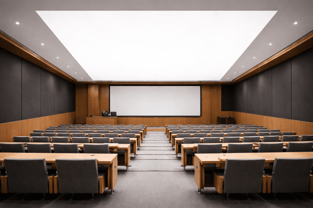 A modern, empty lecture hall with rows of gray chairs and wooden desks faces a large projection screen beneath bright overhead lighting, enhanced by low emission stretch ceiling membranes for improved sustainability.