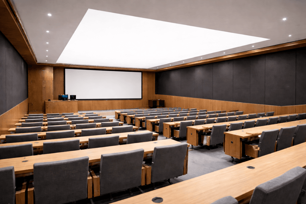A modern, empty lecture hall with rows of wooden desks, gray cushioned chairs, and a large projection screen at the front features wood-paneled walls and bright ceiling lighting, enhanced by Low Emission Stretch Ceiling Membranes.