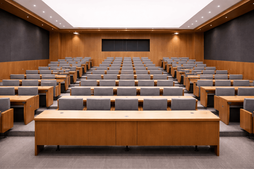A modern, empty lecture hall with wooden desks and rows of gray cushioned chairs faces a large whiteboard at the front. The well-lit room features dark accent walls and showcases design inspired by the Low Emission Stretch Ceiling Membranes Guide.