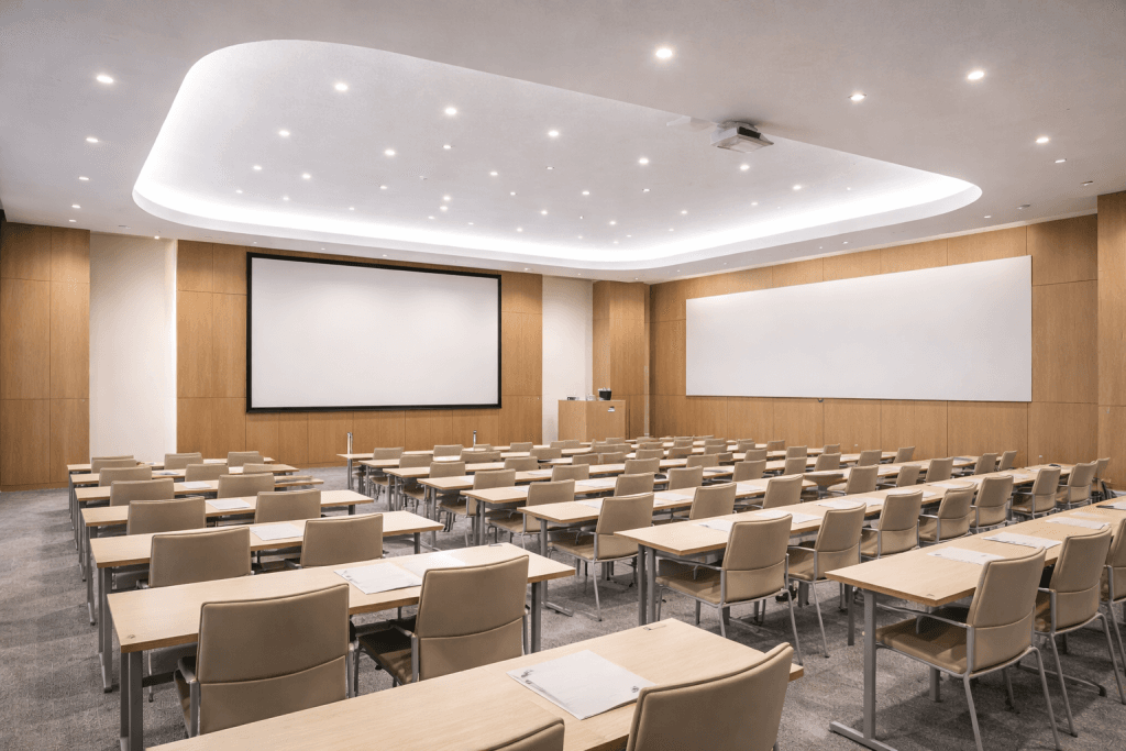 A modern, empty conference room with rows of beige chairs and desks facing a large projection screen and whiteboard, under bright ceiling lights, features light wood paneling designed to support LEED v4.1 Acoustic Panel Credits.