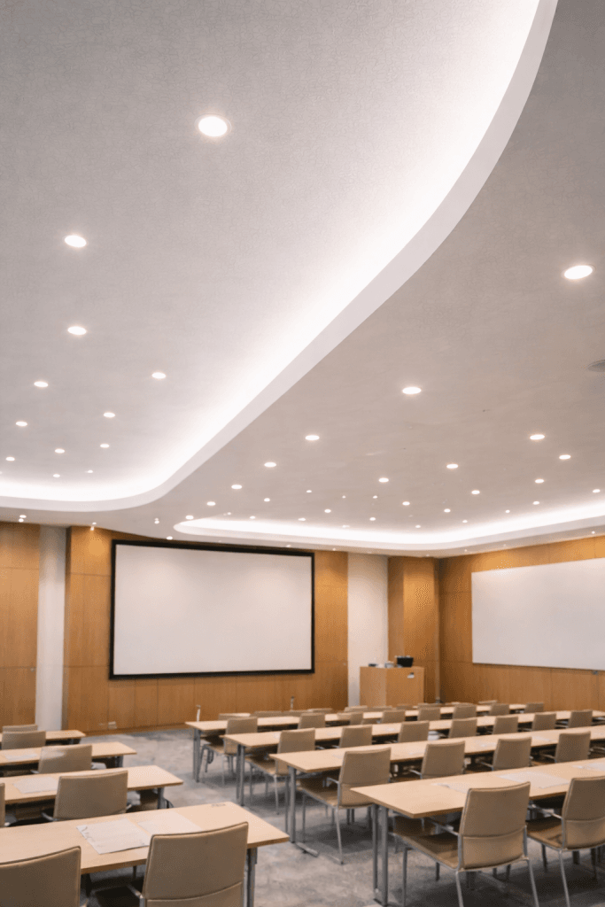 A modern, empty conference room with rows of beige chairs and tables facing two large projection screens, a podium, and a whiteboard. Wooden wall panels feature design elements supporting LEED v4.1 Acoustic Panel Credits and recessed ceiling lights.