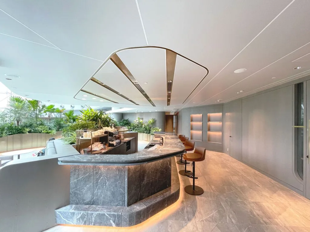 Modern lounge area with a marble counter, tan barstools, indoor plants, and sleek lighting. The space has a curved design, light gray floors, and a bright, airy atmosphere.