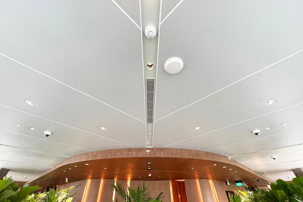 A modern Ligkt™ Stretch Membrane Ceiling features a curved design with recessed lighting, security cameras, and ventilation vents, illuminating a space adorned with green plants and wood-paneled walls.