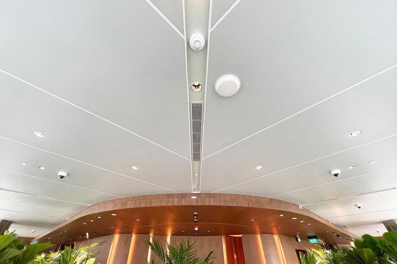 A modern Ligkt™ Stretch Membrane Ceiling with recessed lights, security cameras, and air vents sits above a curved wooden structure. Green plants at the bottom of the image create a fresh interior atmosphere.