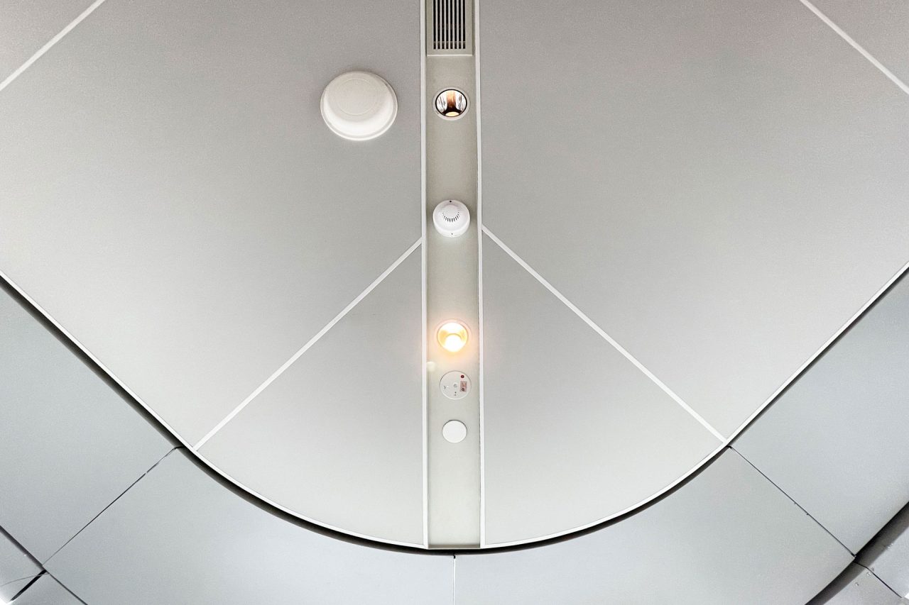 View of a smooth, light gray Ligkt™ Stretch Membrane Ceiling panel with geometric lines, circular vents, a speaker, and two small lights—one illuminated. The design is modern and minimalistic.
