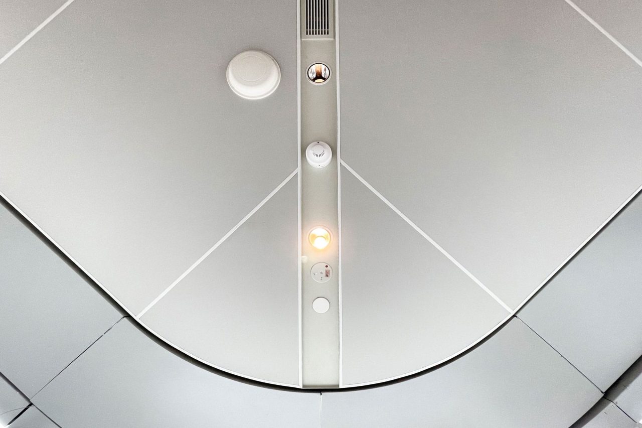 View of a smooth, light gray Ligkt™ Stretch Membrane Ceiling panel with geometric lines, circular vents, a speaker, and two small lights—one illuminated. The design is modern and minimalistic.