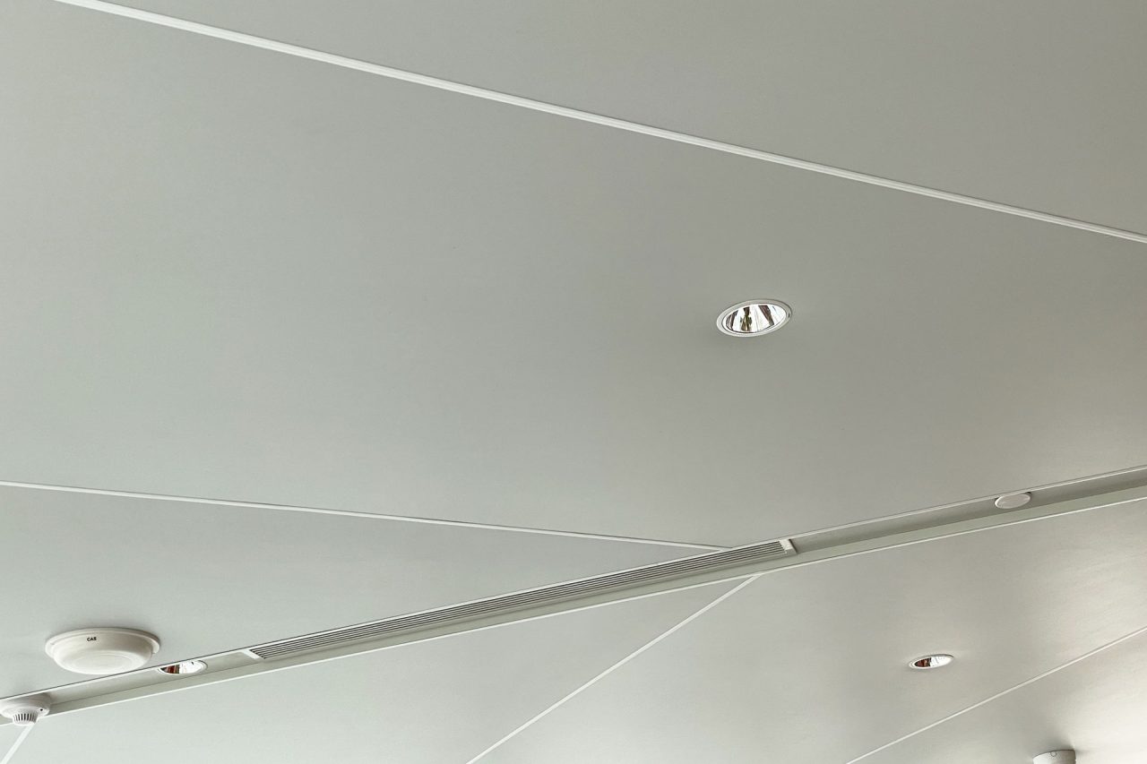 A modern white Ligkt™ Stretch Membrane Ceiling with geometric panel lines, recessed lights, a smoke detector, and a small security camera visible in the corners.