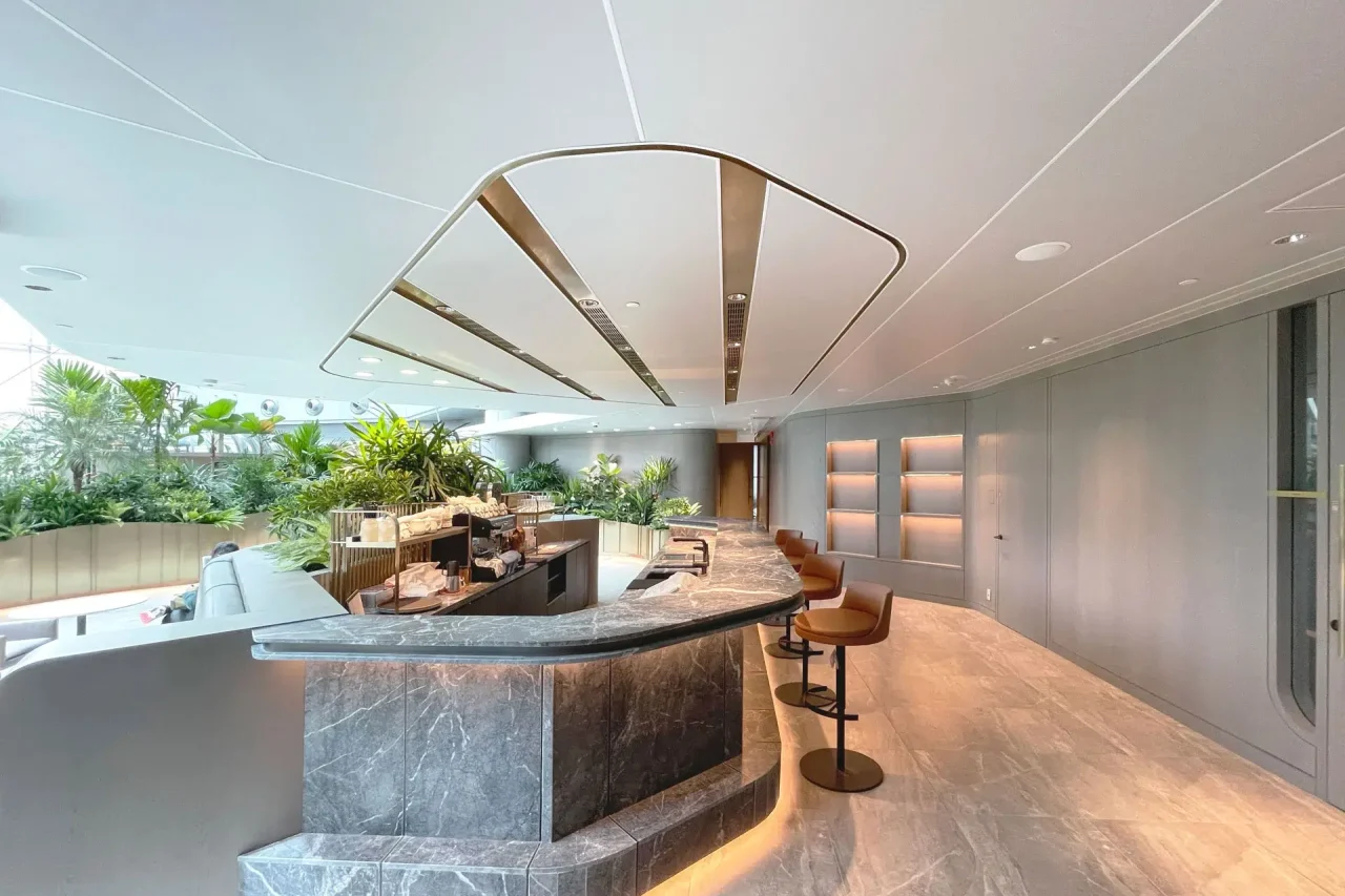 A modern, spacious lounge with a curved marble reception desk, Ligkt™ Stretch Membrane Ceiling, two tan barstools, sleek lighting, and abundant green plants creating a fresh atmosphere. Marble flooring and soft grey walls complete the elegant design.
