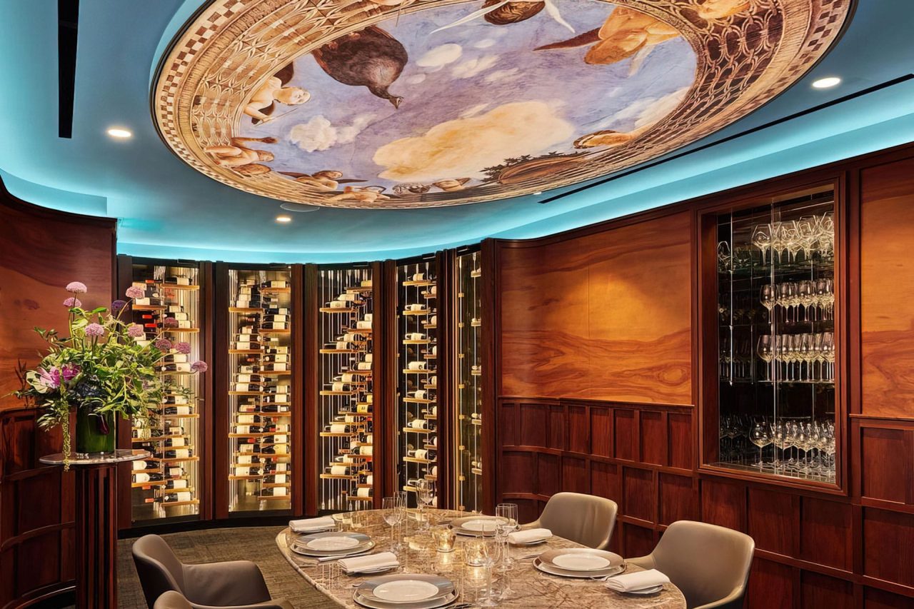 A luxurious dining room with a round table set for six, surrounded by wine racks and wood-paneled walls. The ceiling features a Ligkt™ Stretch Membrane Ceiling with a large, colorful mural of clouds, sky, and classical figures. Modern lighting accents the space.